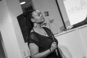Black and white image of a young Black Woman modeling a black dress in a fashion show. She is holding a long stemmed rose.