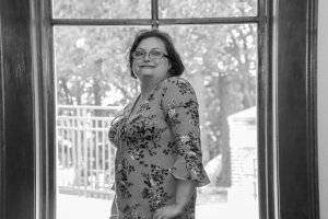 Black and white image of a woman modeling a floral dress in a fashion show. She is standing in front of a large window.